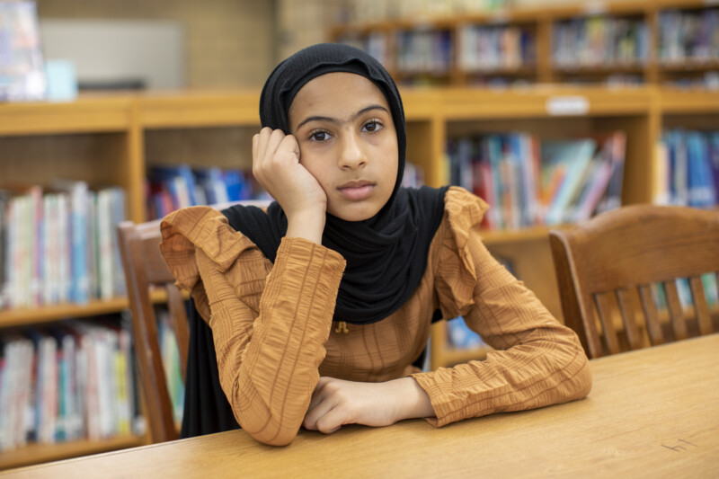A young girl wearing a black hijab and brown top sits at a table in a library, resting her head on her hand and looking thoughtfully at the camera. Rows of books are visible in the background.