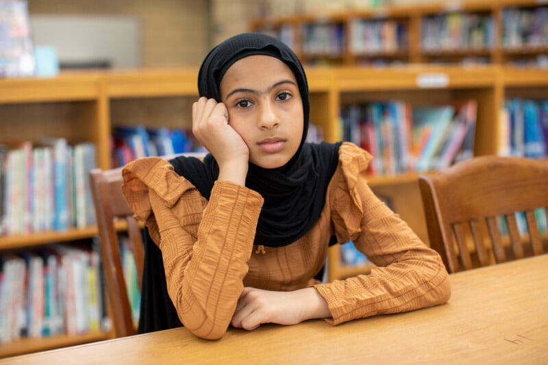 A young girl wearing a black hijab and brown top sits at a table in a library, resting her head on her hand and looking thoughtfully at the camera. Rows of books are visible in the background.