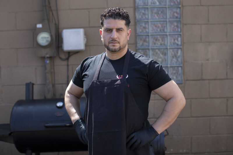 A man wearing a black apron and gloves stands with hands on hips in front of an outdoor smoker grill, against a brick wall with utility boxes and glass blocks.