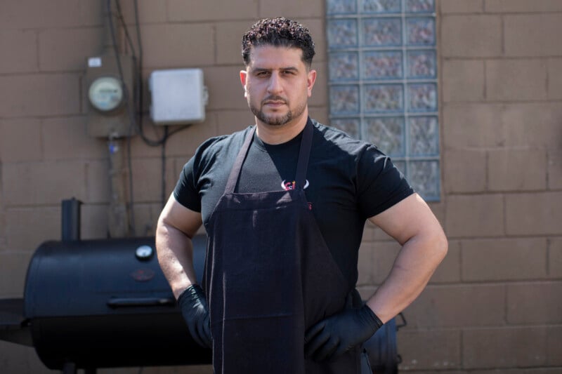 A man wearing a black apron and gloves stands with hands on hips in front of an outdoor smoker grill, against a brick wall with utility boxes and glass blocks.