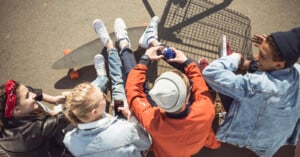 Four young people sit together on the ground outdoors, some wearing denim jackets and sneakers, with a skateboard and a shopping cart nearby, chatting and enjoying drinks in the sunlight.