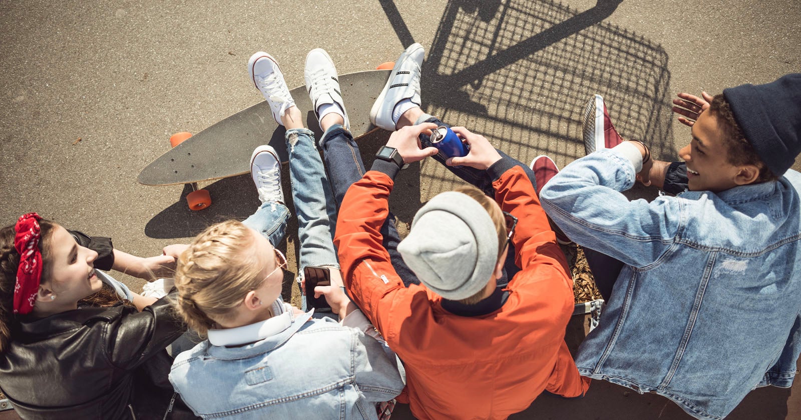 Four young people sit together on the ground outdoors, some wearing denim jackets and sneakers, with a skateboard and a shopping cart nearby, chatting and enjoying drinks in the sunlight.