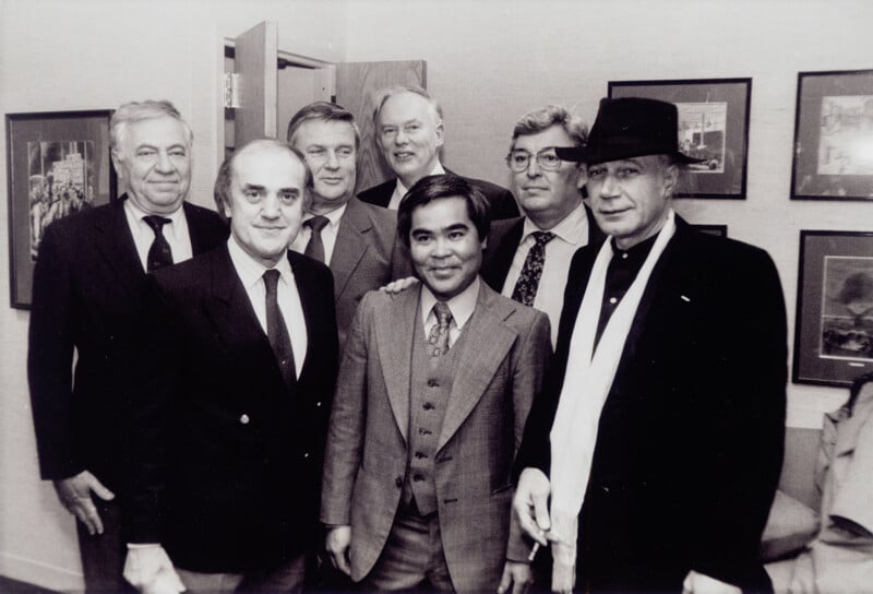 A group of seven men in suits and ties, some smiling, pose together in an office with framed pictures on the wall. One man on the right wears a hat and scarf. The photo is in black and white.