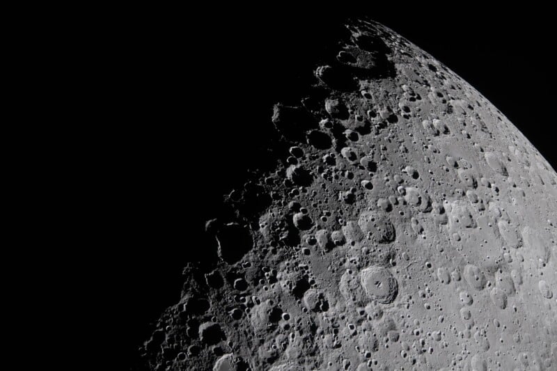 A detailed close-up of the moon’s surface shows numerous craters and rugged terrain. The lighting creates strong shadows, highlighting the texture and depth of the lunar landscape against a black background.