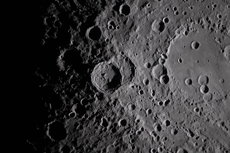 A detailed, high-resolution image of the Moon’s surface showing numerous craters of various sizes, with shadows accentuating their depth and rugged texture. The left side is partially in darkness.