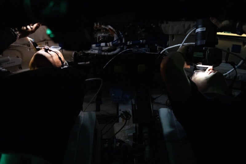 Two people work in a dimly lit, cramped space surrounded by wires and equipment, each using a flashlight to illuminate their tasks, creating a focused and shadowy atmosphere.