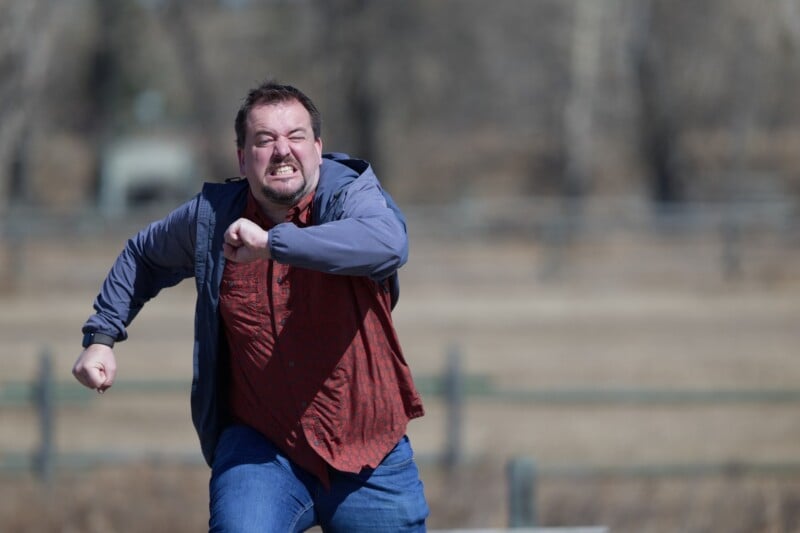 A man with a tense expression is running outdoors. He wears a red shirt and blue jacket, with his arms bent and fists clenched. The background is blurred with bare trees and a fence.