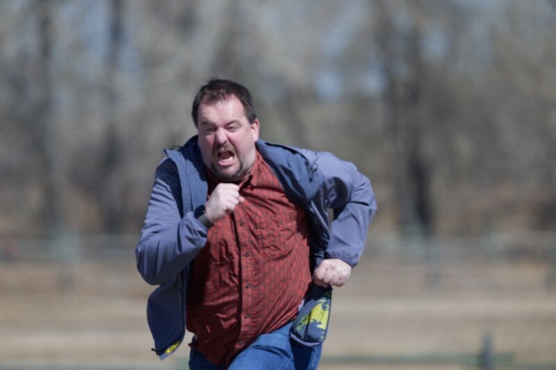 A man wearing a red shirt and gray jacket is running outdoors with a determined expression. The background is blurred, showing trees and a fence.