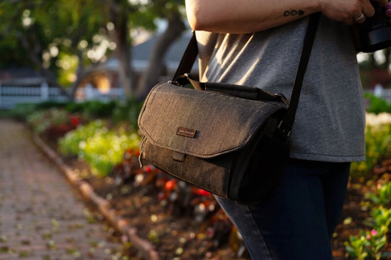 A person wearing a gray shirt carries a black camera bag with a shoulder strap while standing on a garden path with flowers and sunlight in the background.