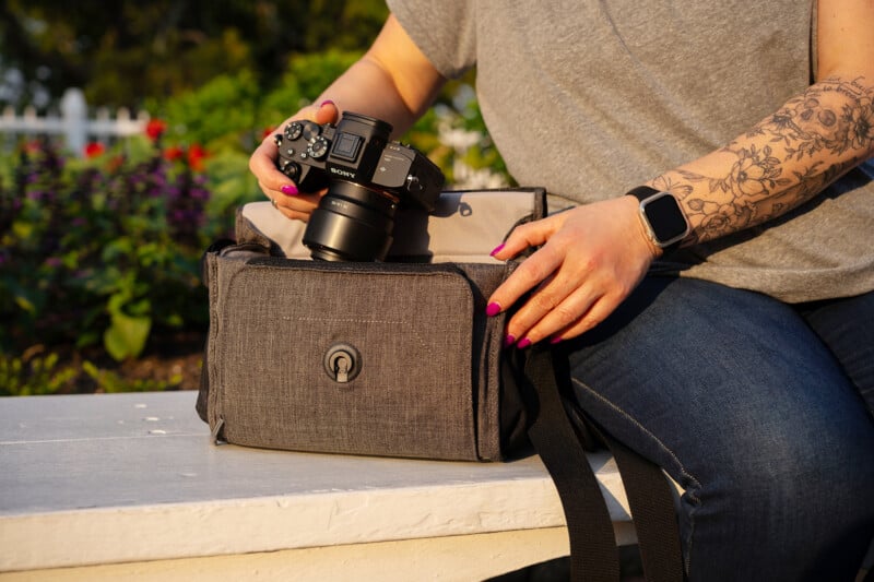 A person with a floral tattoo and pink nail polish puts a black Sony camera into a gray camera bag while sitting on a white bench outdoors.