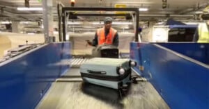 A worker in a safety vest loads luggage onto a conveyor belt in an airport baggage handling area, with suitcases moving toward the camera.