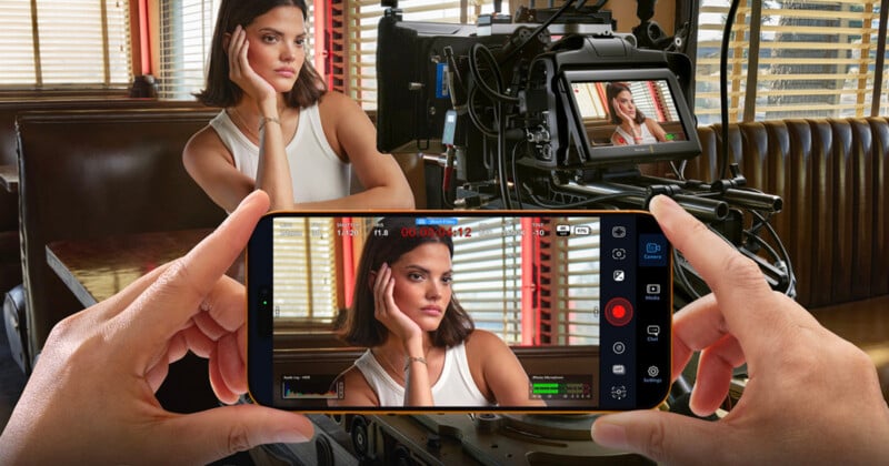A woman sits in a diner booth, resting her head on her hand, while being filmed by a professional camera and a smartphone held in the foreground, both screens showing her pose.