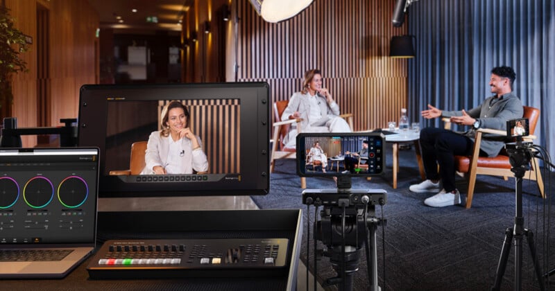 A professional video interview setup shows two people seated and talking, surrounded by cameras, studio lights, monitors, and video production equipment in a modern room with wood paneling and curtains.