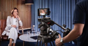 A woman sits in a chair, smiling, during an interview being filmed by a camera operator. She faces the camera, with a table holding glasses and a water carafe in front of her. A phone is attached to the camera as a monitor.