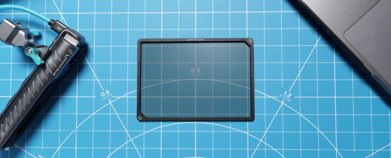 A rectangular camera filter labeled "0.9 3 STOP" lies on a blue grid cutting mat, with a tripod grip on the left and a laptop on the right.
