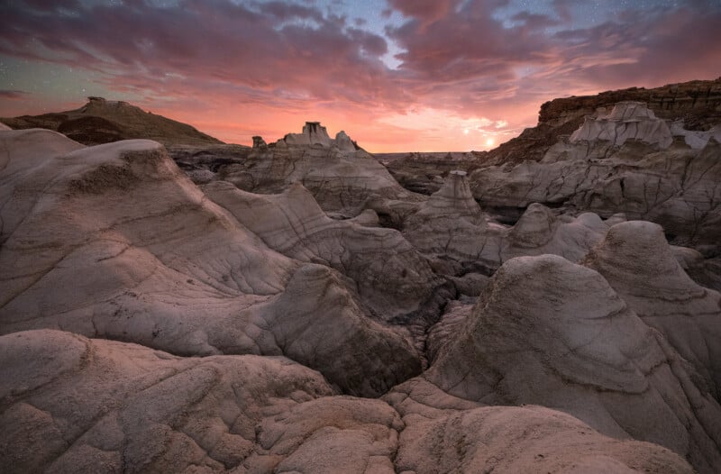 Jagged rock formations and eroded badlands under a dramatic, colorful sunset sky with shades of pink, orange, and purple, creating a striking and otherworldly desert landscape.