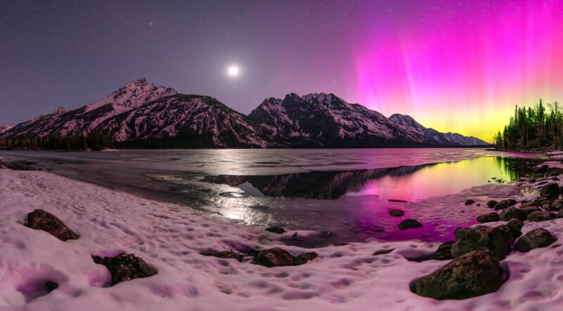 Snow-covered mountains and a partially frozen lake under a starry sky with the moon. Vivid pink, yellow, and green aurora lights illuminate the sky and reflect on the water. Pine trees line the shore.