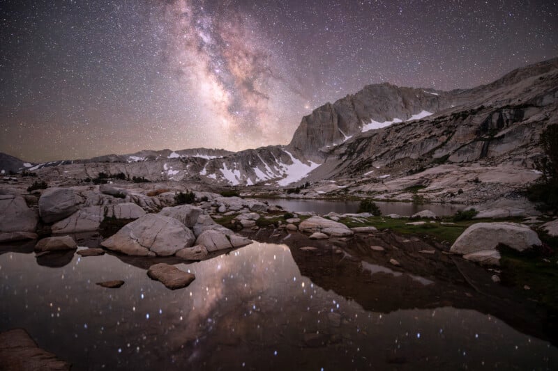 The Milky Way shines brightly in a star-filled night sky above snow-dusted mountains, with the stars and peaks reflected in a calm, rocky alpine lake.