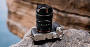 A Sony Alpha camera with a large lens sits on a rocky surface outdoors, with blurred rocks and water in the background. The camera’s top dials and lens details are clearly visible.