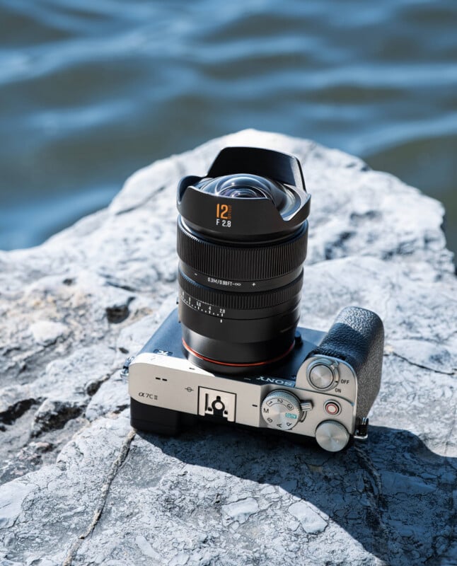A silver Sony Alpha camera with a wide-angle 12mm f/2.8 lens rests on a textured gray rock near a body of water under bright sunlight.