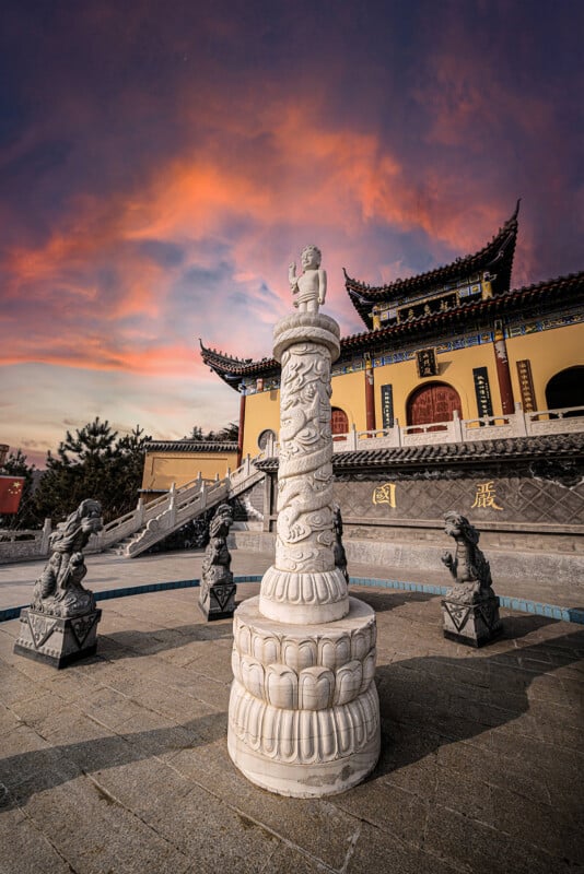A tall white stone pillar with intricate dragon carvings stands in front of a traditional Chinese temple, surrounded by dark statues, under a vibrant sunset sky with pink and orange clouds.