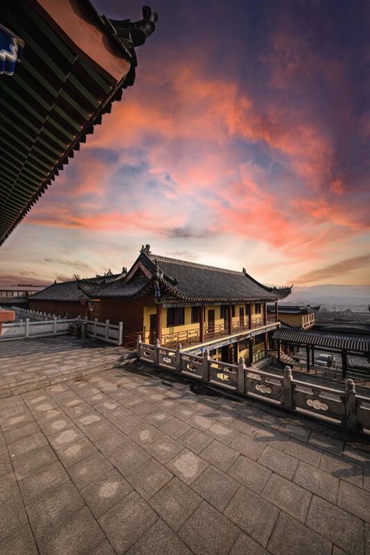 Traditional East Asian building with ornate roof tiles and wooden railings at sunset, vibrant pink and orange clouds filling the sky, and stone courtyard in the foreground.