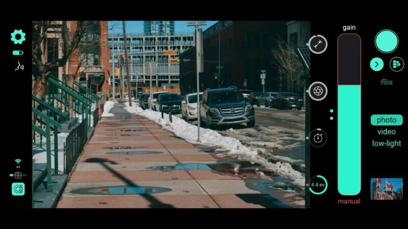 A city sidewalk with meltwater puddles and snow piles is shown. Parked cars line the street. On-screen camera settings appear on the right, suggesting a photo or video is being taken.