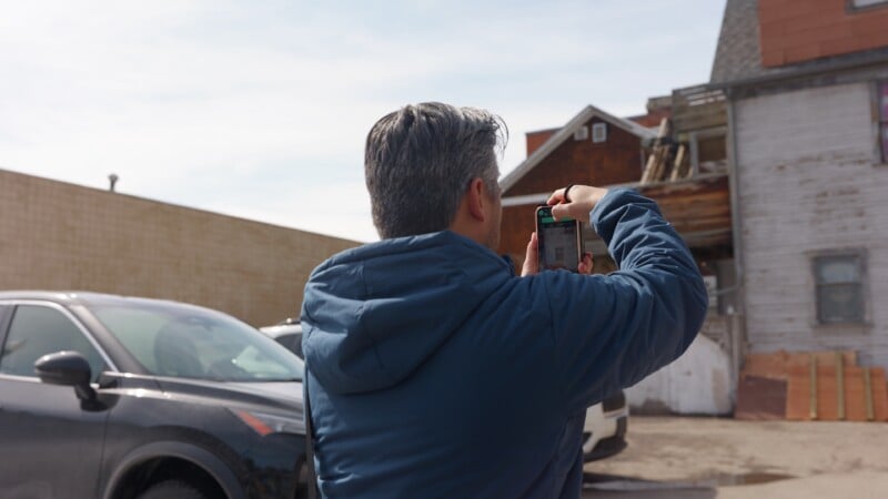 A person with gray hair in a blue jacket takes a photo of an old, weathered house using a smartphone in a parking lot with a black car nearby.