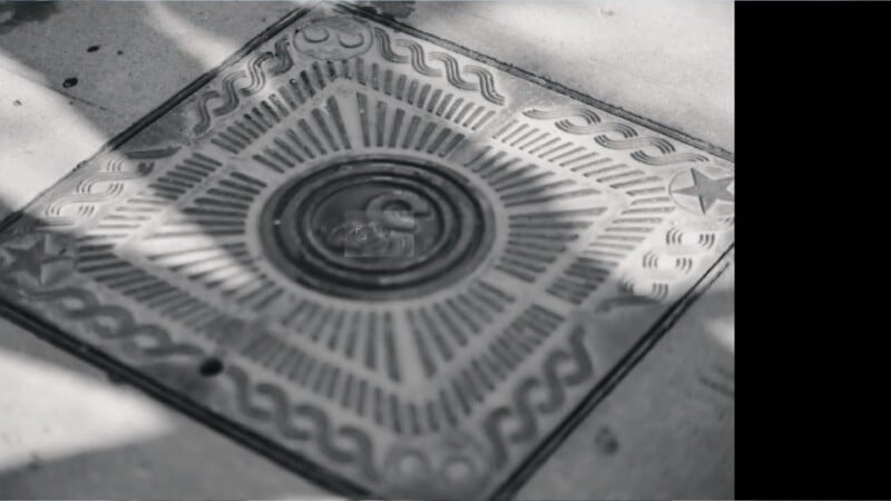 A decorative metal manhole cover with geometric and wave patterns is set in a concrete sidewalk. Shadows and light fall across the cover, which features a central circle and star shapes in the corners.