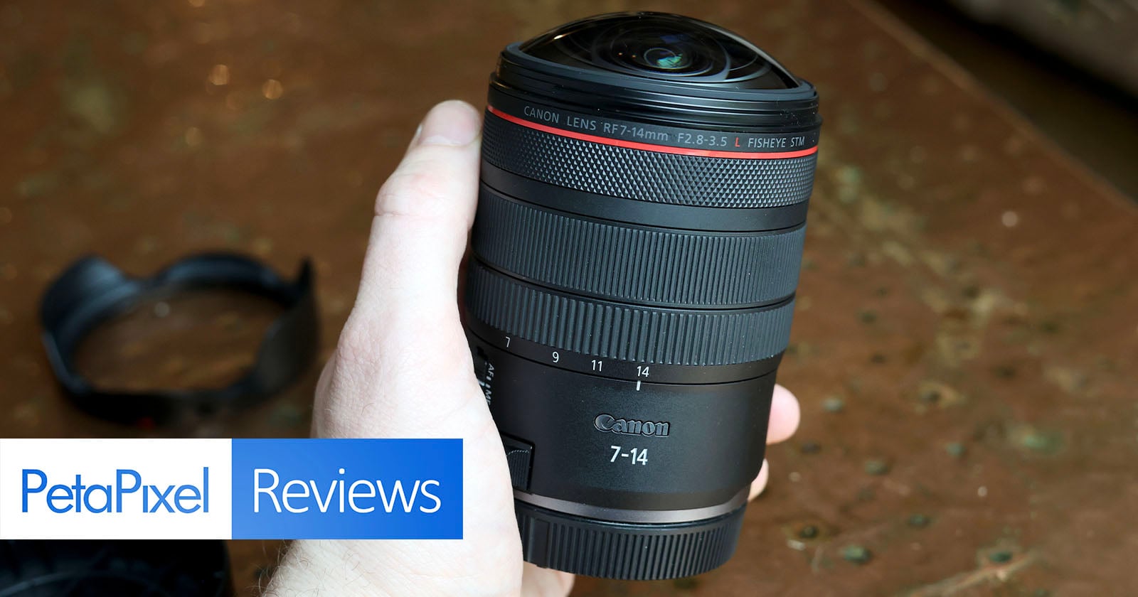 A hand holds a Canon RF 14-35mm F2.8 L IS USM camera lens, with a lens cap nearby. The image includes a "PetaPixel Reviews" graphic in the bottom left corner.
