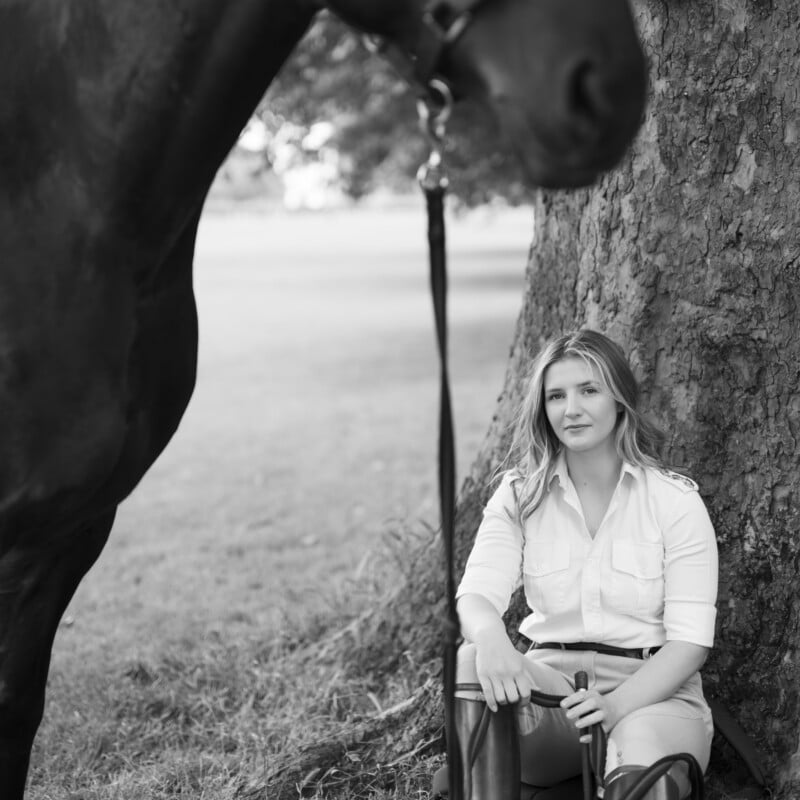 A woman in equestrian attire sits under a tree, holding riding gear, with a horse standing beside her. The scene is outdoors in a grassy area, captured in black and white.