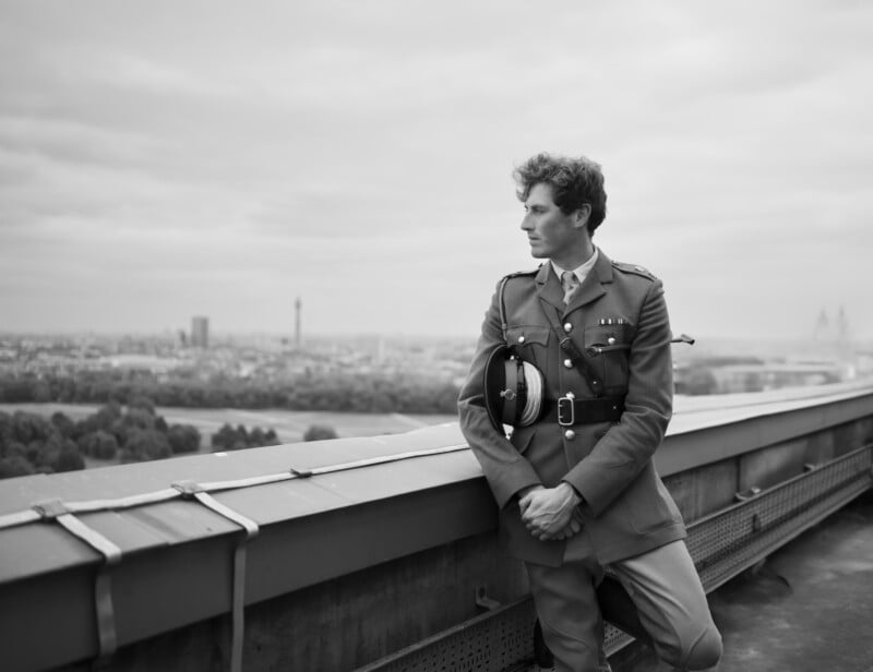 A man in a military uniform stands on a rooftop, leaning against a barrier and holding his hat. The city skyline and trees are visible in the distance under a cloudy sky.