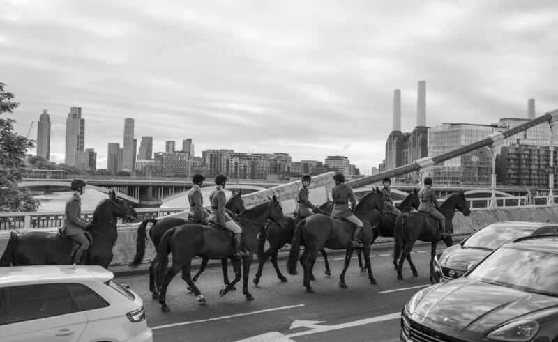 A group of mounted police officers ride horses along a busy city street by the river, with modern skyscrapers and industrial buildings in the background. Cars are visible in the foreground. The image is in black and white.