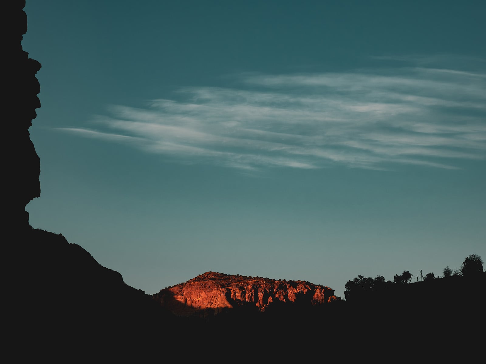 A dramatic landscape shows a sunlit red rock formation glowing at sunset, surrounded by dark silhouettes of cliffs and sparse vegetation under a dusky blue sky with wispy white clouds.