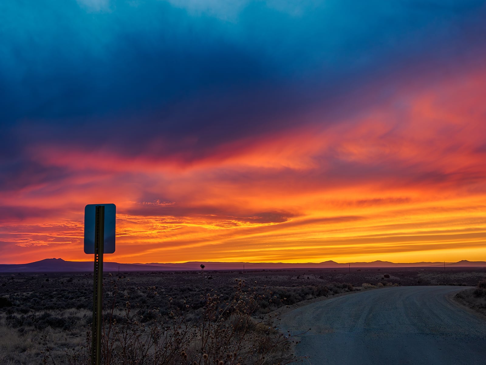 A vivid sunset paints the sky with orange, yellow, pink, and blue hues above a rural landscape. A dirt road curves to the right, with a roadside sign silhouetted in the foreground and distant mountains on the horizon.