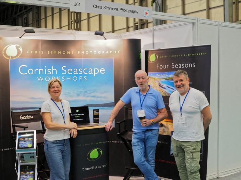 Three people stand and smile at a photography exhibition booth for "Chris Simmons Photography," featuring banners for Cornish Seascape Workshops and Four Seasons. The booth includes photos, brochures, and a display counter.