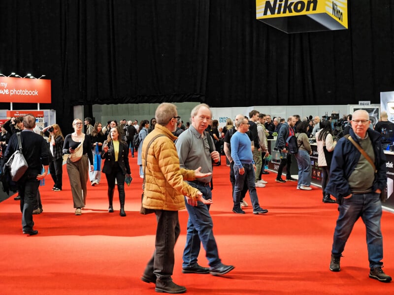 People walk around a busy exhibition hall with a red carpet. Various stalls and company signs, including Nikon, are visible in the background. The atmosphere appears lively, with attendees conversing and exploring booths.