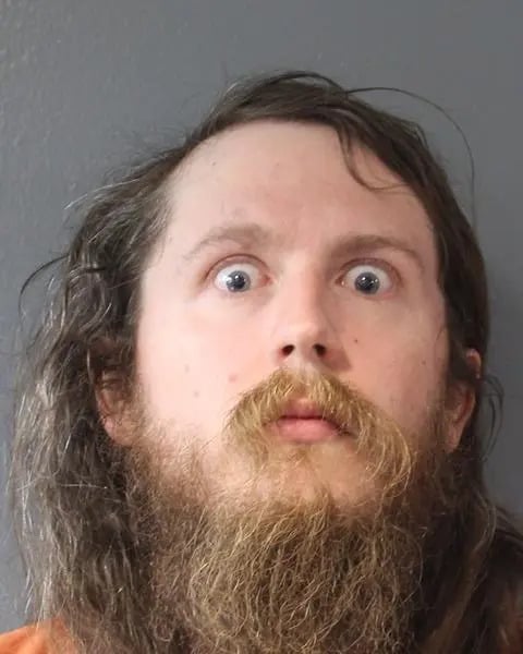 A man with long brown hair and a thick beard stares wide-eyed at the camera with a surprised expression, standing against a plain gray background.