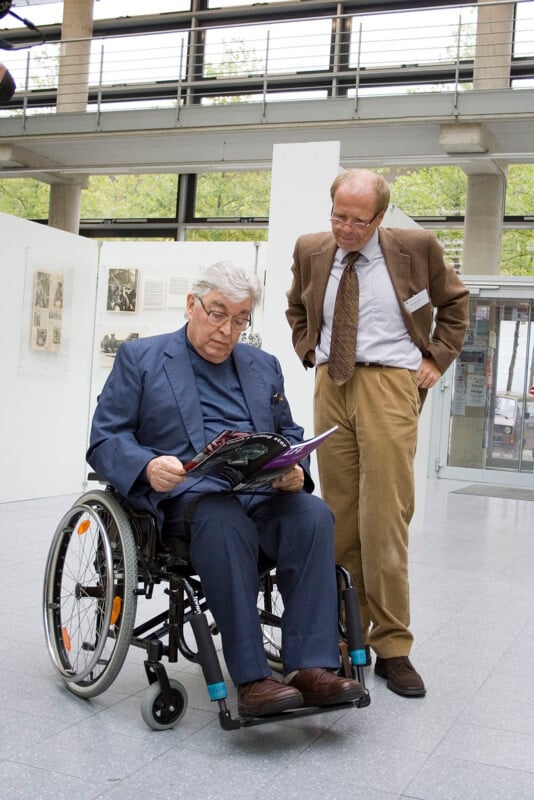 An older man in a wheelchair reads a book while another man stands beside him, leaning slightly forward and looking at the book. They are indoors in a modern, well-lit building with large windows.
