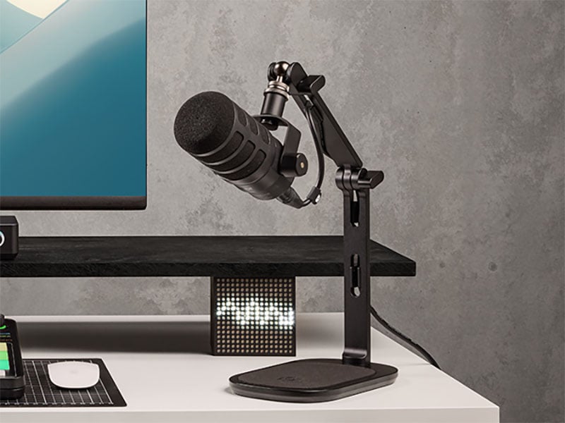 A black microphone on an adjustable arm stand is positioned on a white desk next to a monitor. A digital pixel display and keyboard are also visible on the desk in a modern workspace.