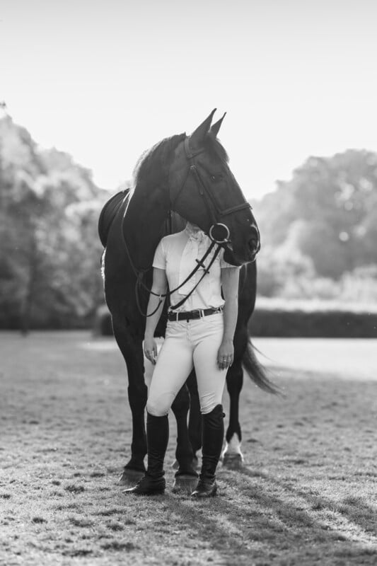 A woman in riding gear stands in a field with a black horse behind her, positioned so the horse’s head covers her own, creating an amusing optical illusion. The scene is in black and white.