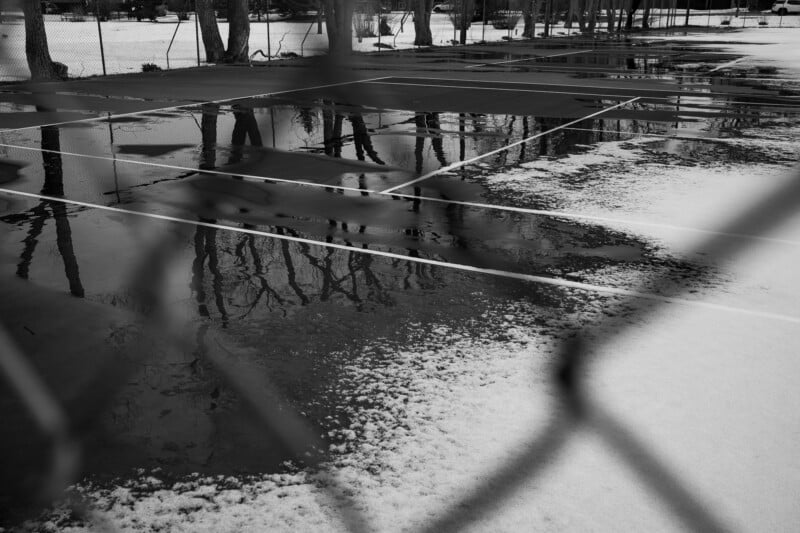 A snow-covered tennis court with patches of melted ice and reflections of trees in the puddles, seen through a chain-link fence on a cold winter day.