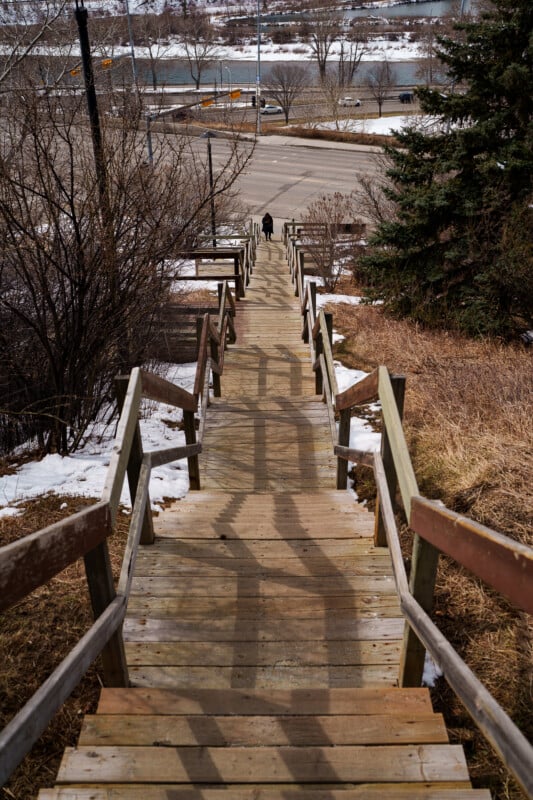 A long wooden staircase descends toward a road, flanked by leafless trees and patches of snow. A lone person is walking at the bottom of the stairs near a river in the background.