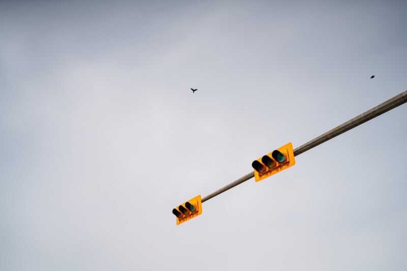 A yellow traffic light hangs horizontally against a pale, cloudy sky. Two birds are flying in the distance near the upper center of the image.