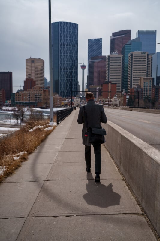 A man in a gray coat walks alone on a city sidewalk toward tall downtown buildings, carrying a black shoulder bag. The day is overcast, and there are patches of snow on the ground.