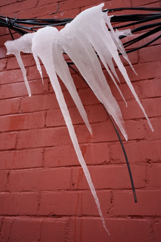 Large icicles hang from black cables against a red brick wall, with the icicles pointing downward in various lengths and shapes.