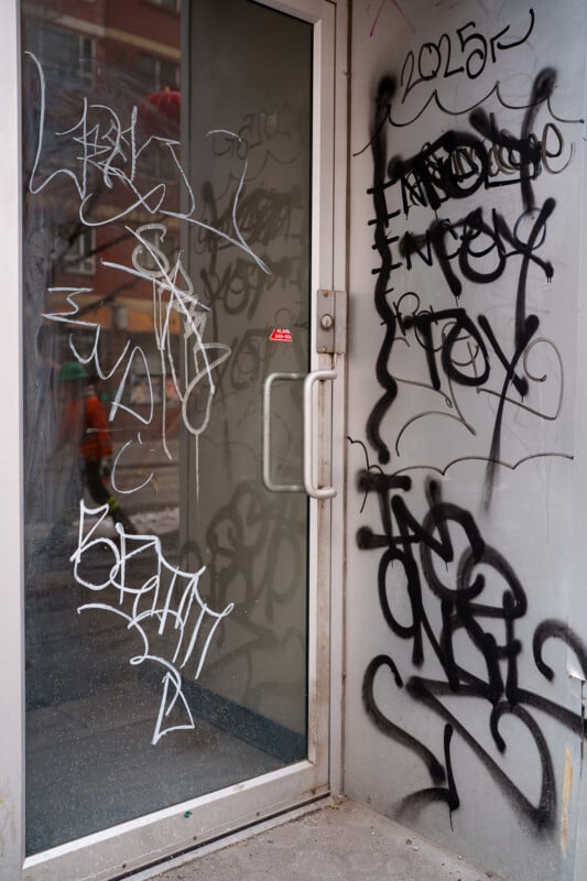 A glass door and surrounding wall are covered with various graffiti tags and scribbles in black and white spray paint. The door has a metal frame and handle, and the ground is concrete.