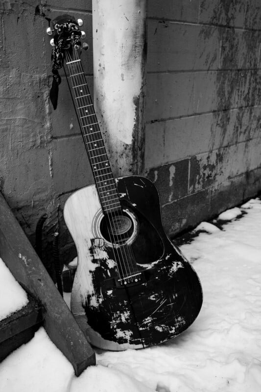 A worn, black-and-white acoustic guitar with chipped paint leans against a concrete wall near snowy steps in an outdoor setting.