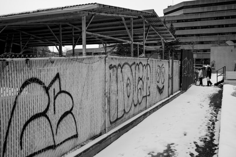 A snow-covered path runs beside a chain-link fence with graffiti. Two people stand near the gate at the end of the path. A large, windowed building and a metal-roofed structure are in the background.