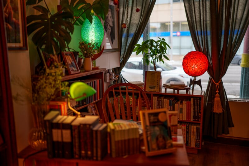 A cozy reading nook with wooden shelves full of books, a wooden chair, plants, and colorful glowing lamps by a window with dark curtains. Some books and framed photos are displayed on the shelves and table.